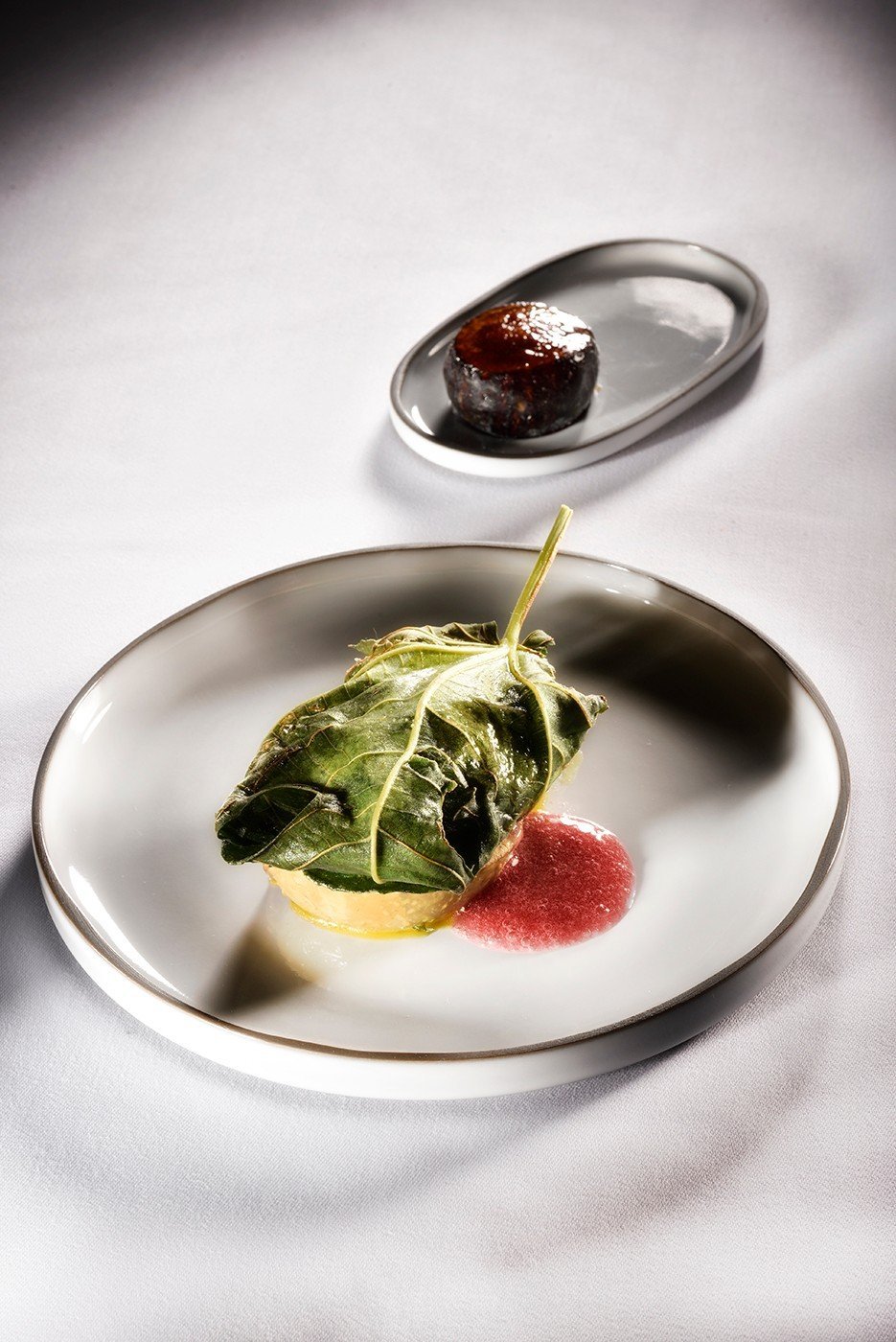 Assiette gastronomique : une préparation enveloppée dans une grande feuille verte, un trait de coulis rouge, une mignardise en arrière-plan